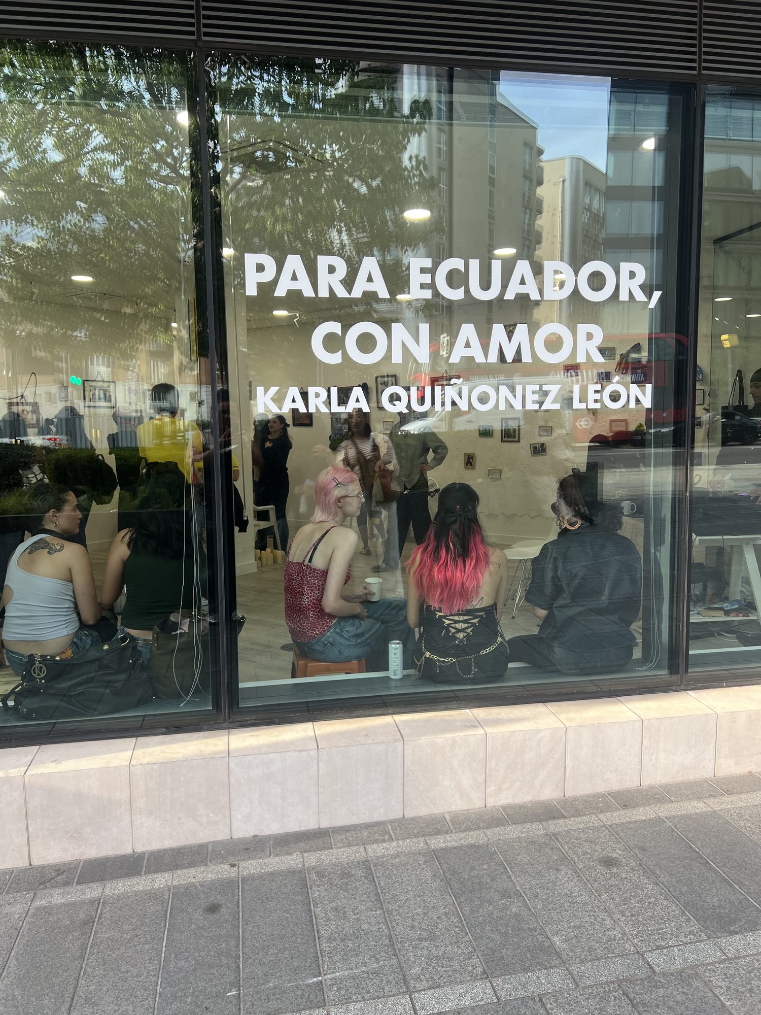 Storefront window reading 'Para Ecuador, Con Amor — Karla Quiñonez León' with visitors gathered inside the exhibition